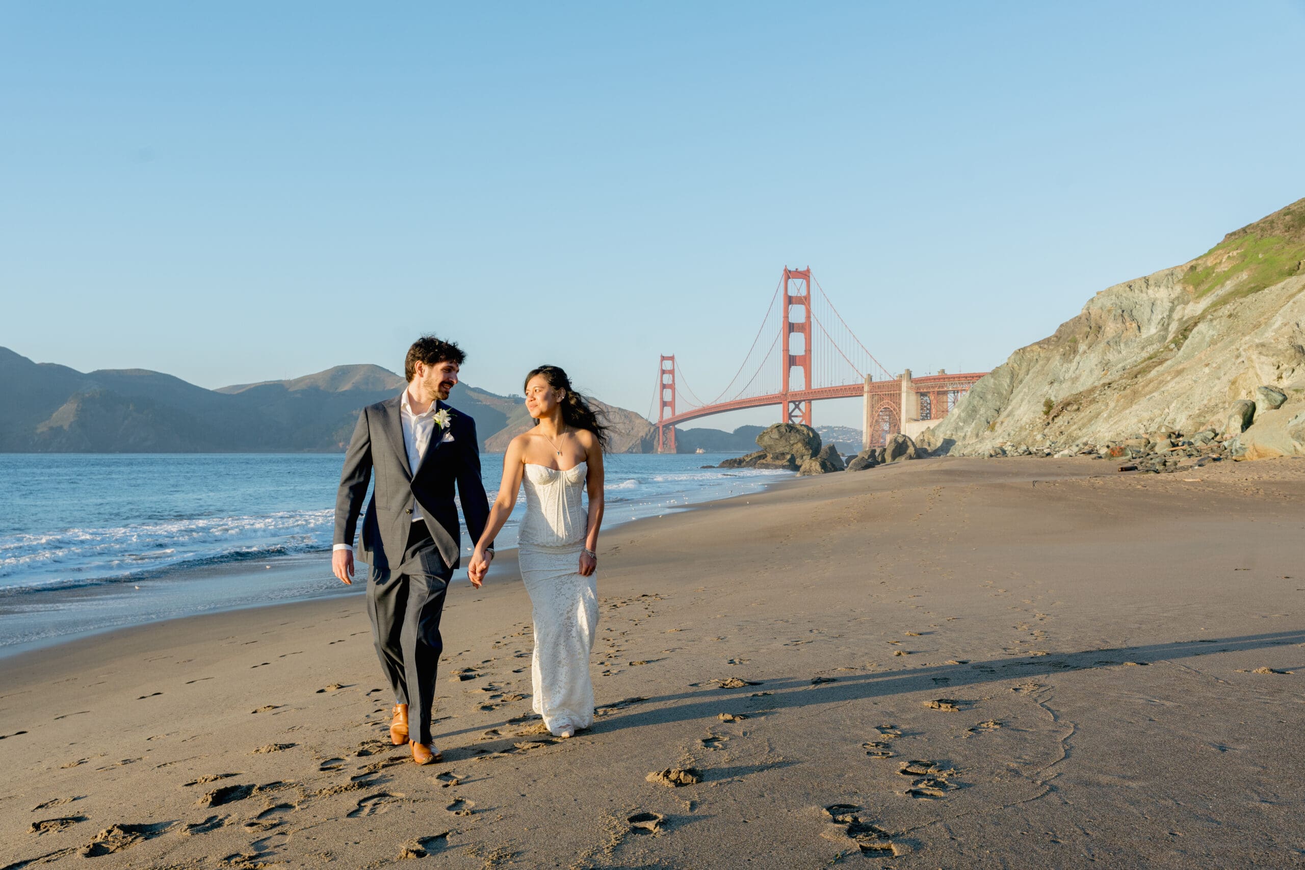 Bride and groom walking hand-in-hand along the shore at Marshall's Beach during a sunset elopement with the Golden Gate Bridge in the background.